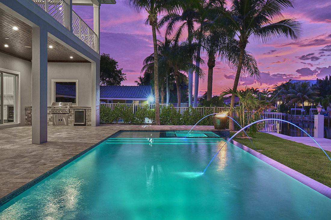 Backyard pool with deck jets and lighting at sunset with palm trees, outdoor kitchen and covered lanai